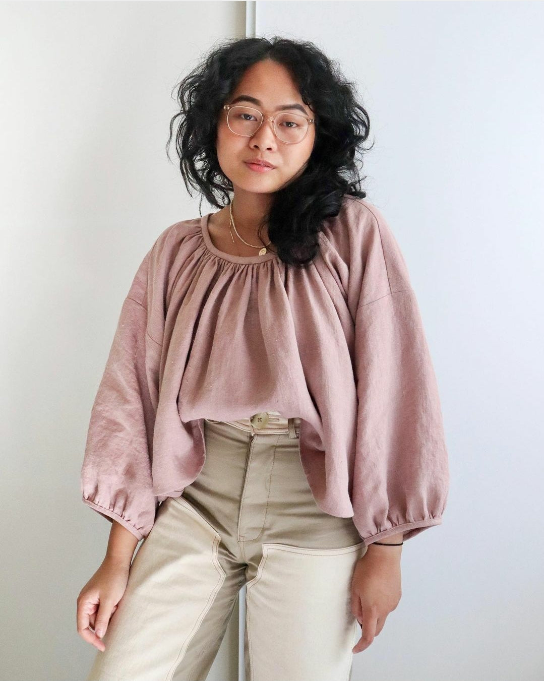 A person wearing a pinkPhotinia top with a gathered neckline and long sleeves, paired with beige pants.